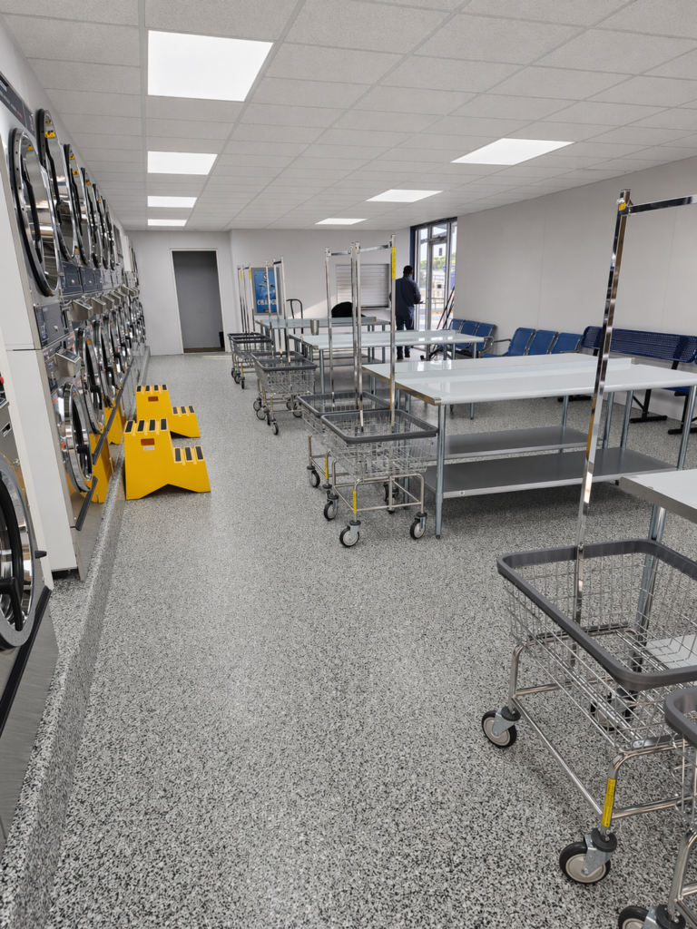 Interior of a laundromat with stacked washers and dryers, rolling laundry carts, folding tables, and blue seating along the wall
